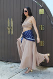 Woman in a beige top and long skirt with a blue polka dot wrap standing in front of a large wooden door.