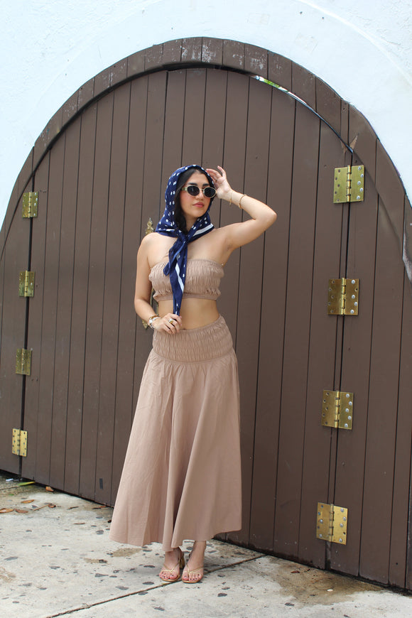 Woman in a beige dress with a blue scarf standing in front of a wooden door.